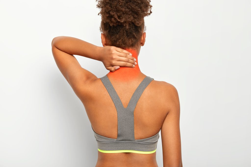 back view dark skinned curly woman touches neck feels pain needs massage suffers from muscle injury wears grey top isolated white wall KPM Physiotherapy
